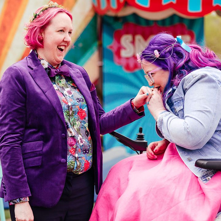 Bride in purple velvet blazer and black suit trousers with purple bowtie, pink patterned pocket square and colourful patterned shirt smiling and laughing with bride in wheelchair wearing personalised denim jacket, pink wedding dress, blue tights and gemstone bridal crown 