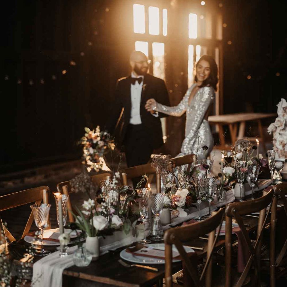 Bride in silver sparkly wedding dress and groom in classic black tuxedo with black bow tie walking by rustic luxe wedding tablescape 