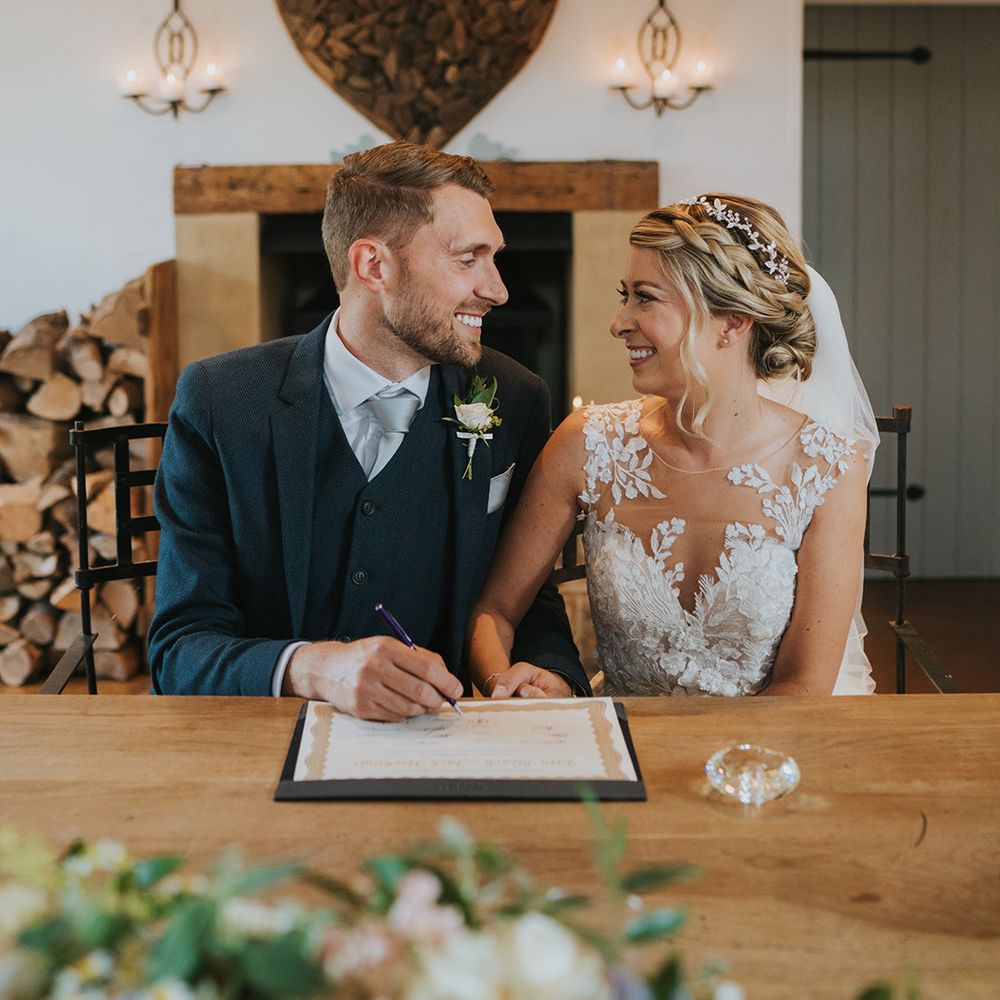 Groom in dark navy three piece suit with silver grey tie smiles at the bride as they sign the wedding register together 
