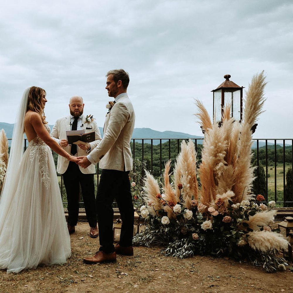 Bride in white embroidered tulle dress holds grooms hands at Italian wedding ceremony surrounded by pampas grass and white, orange and pink rose decor
