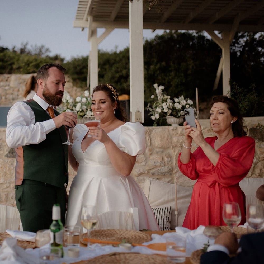 Bride in white satin wedding dress cheersing the groom in a dark green suit with a cocktails and red wine at private villa wedding