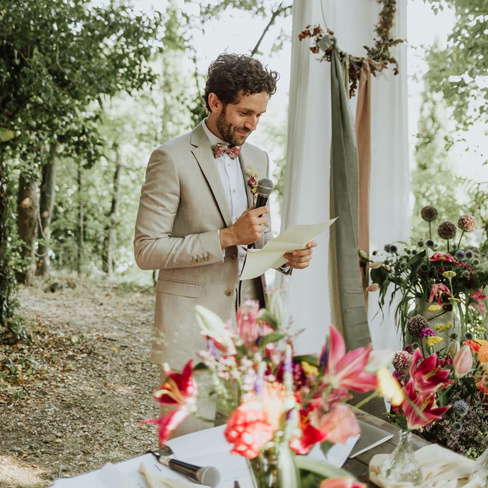 Groom in griege suit and colourful bow tie reads his written words to the bride during Italian woodland wedding ceremony
