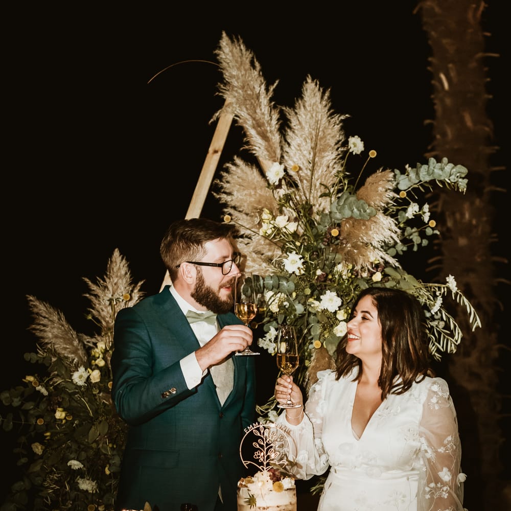 Bride and groom toasting in front of their single tier semi naked wedding cake and cupcake table 