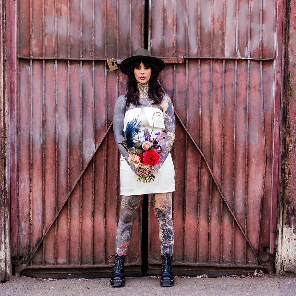 Tattooed bride in a short wedding dress with Dr Marten boots and fedora hat holding a colourful wedding bouquet 