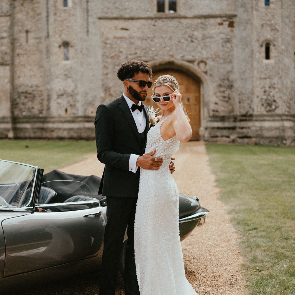 Bride in an appliqué wedding dress, peal headband and white sunglasses standing outside Pentney Abbey with her husband in a tuxedo 