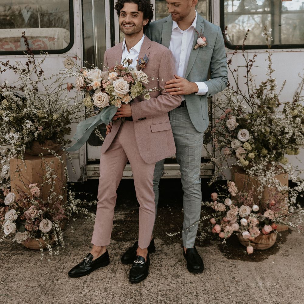 Grooms stand outdoors in front of floral installations whilst smiling and groom holds arm of partner