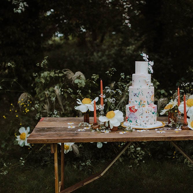 four tier wedding cake with flower illustration resting on a wooden table with wildflower decor 