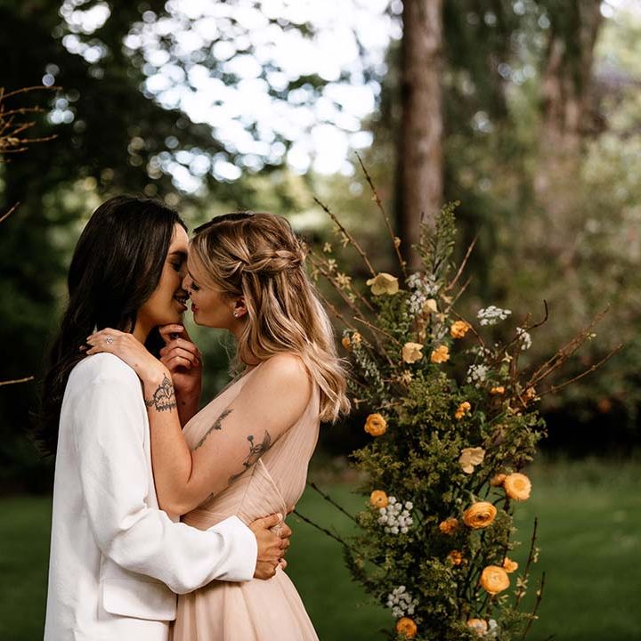 Bride with tattoos in a blush pink wedding dress kissing her bride at the altar in a white trouser suit 