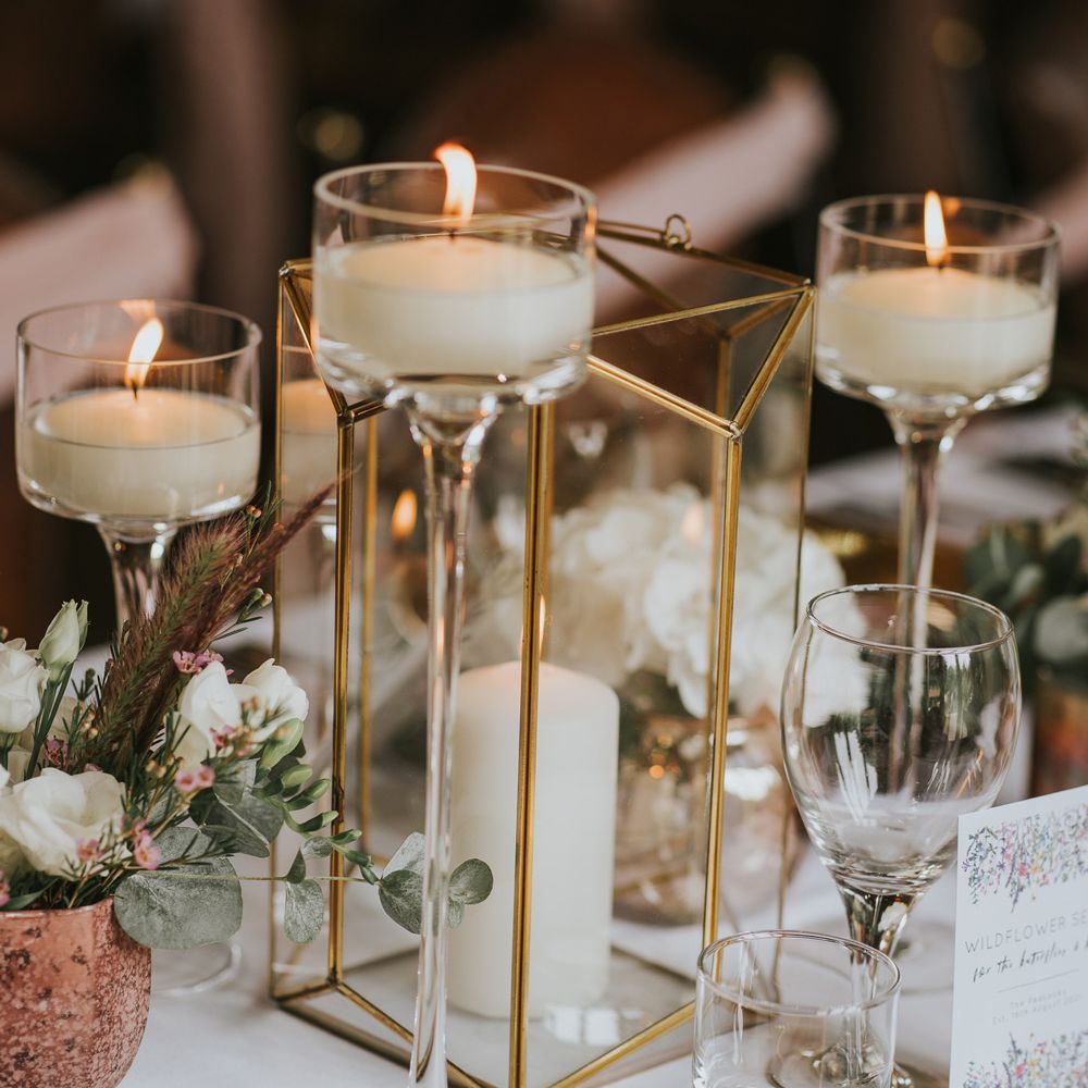 Gold candle box with pillar candle and long stem glass candle holders on white tablecloth next to gold plates and glassware at Tythe Barn wedding with barn wedding flowers