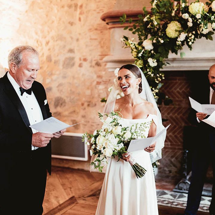Bride stands at the front of the aisle wearing off the shoulder gown and carrying white floral bouquet