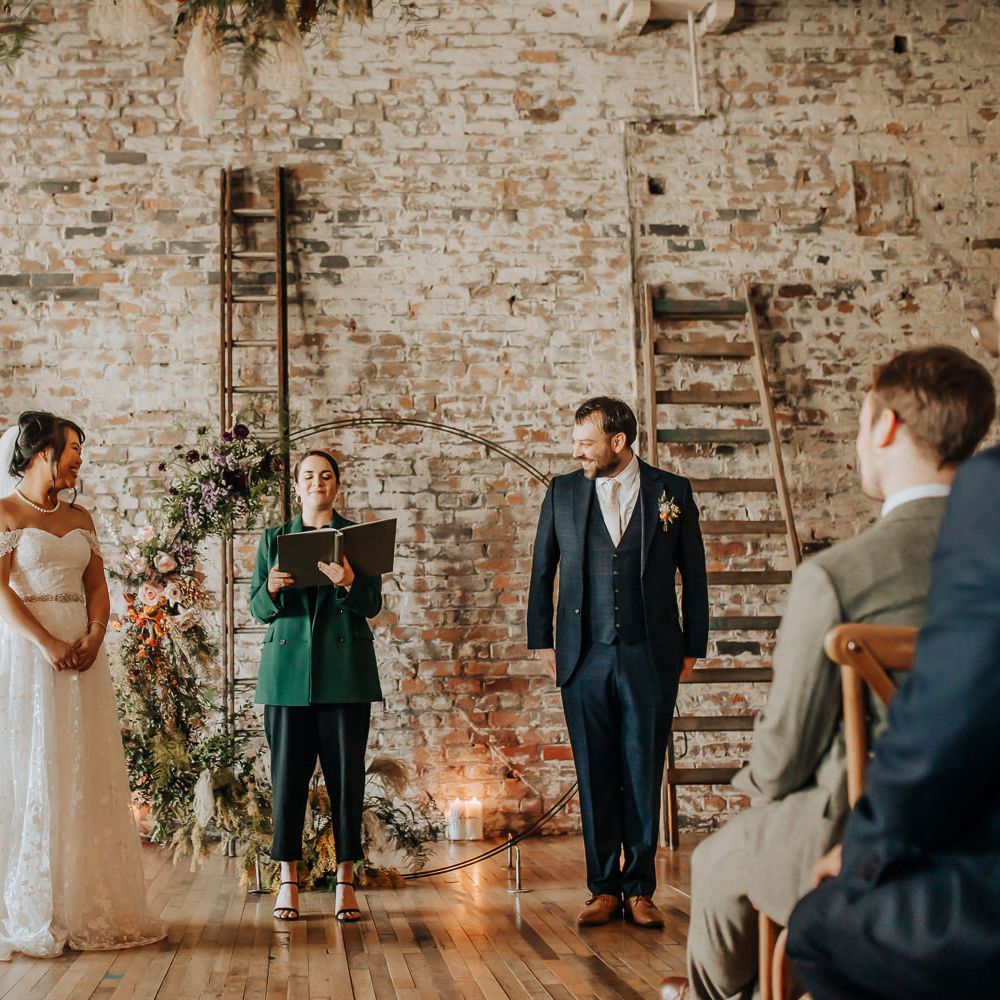 Celebrant in green blazer reads whilst bride in lace bardot wedding dress, pearl necklace and veil stands with groom in blue three piece suit and pink tie during wedding ceremony