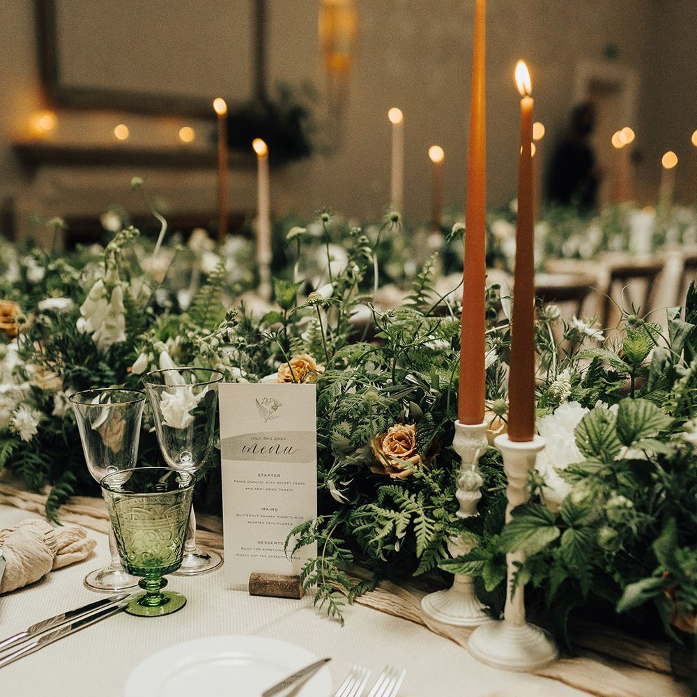 Elegant wedding table decor and place setting with white candlestick holders, brown taper candles and green foliage wedding flowers 
