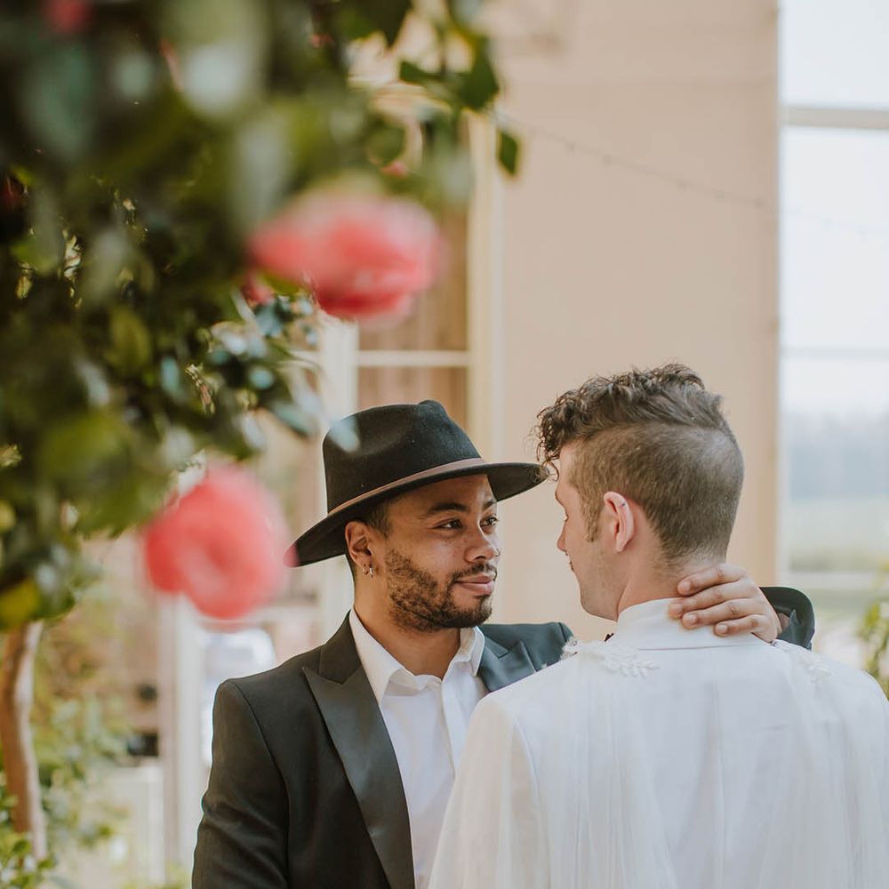 Groom in a black suit and fedora hat embracing his partner in a white groom suit and cape