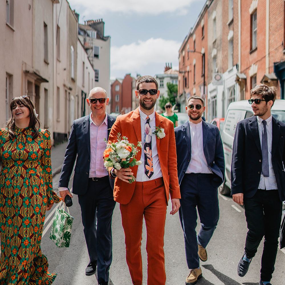 Groom in sunglasses and orange Paul Smith linen suit holds bridal bouquet and walks alongside wedding party in the streets of Bristol 