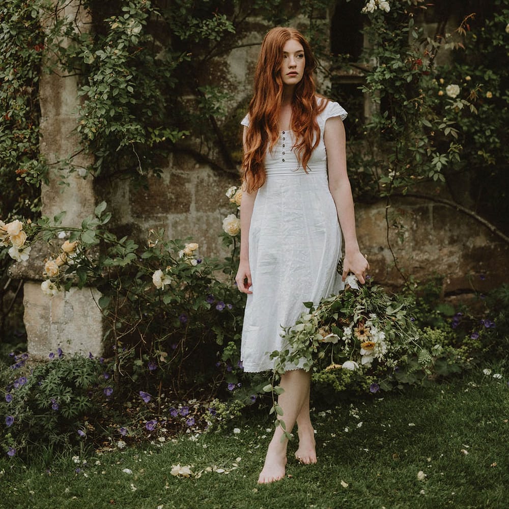 Barefoot bride with long wavy red hair in a linen midi wedding dress holding a green foliage bridal bouquet 