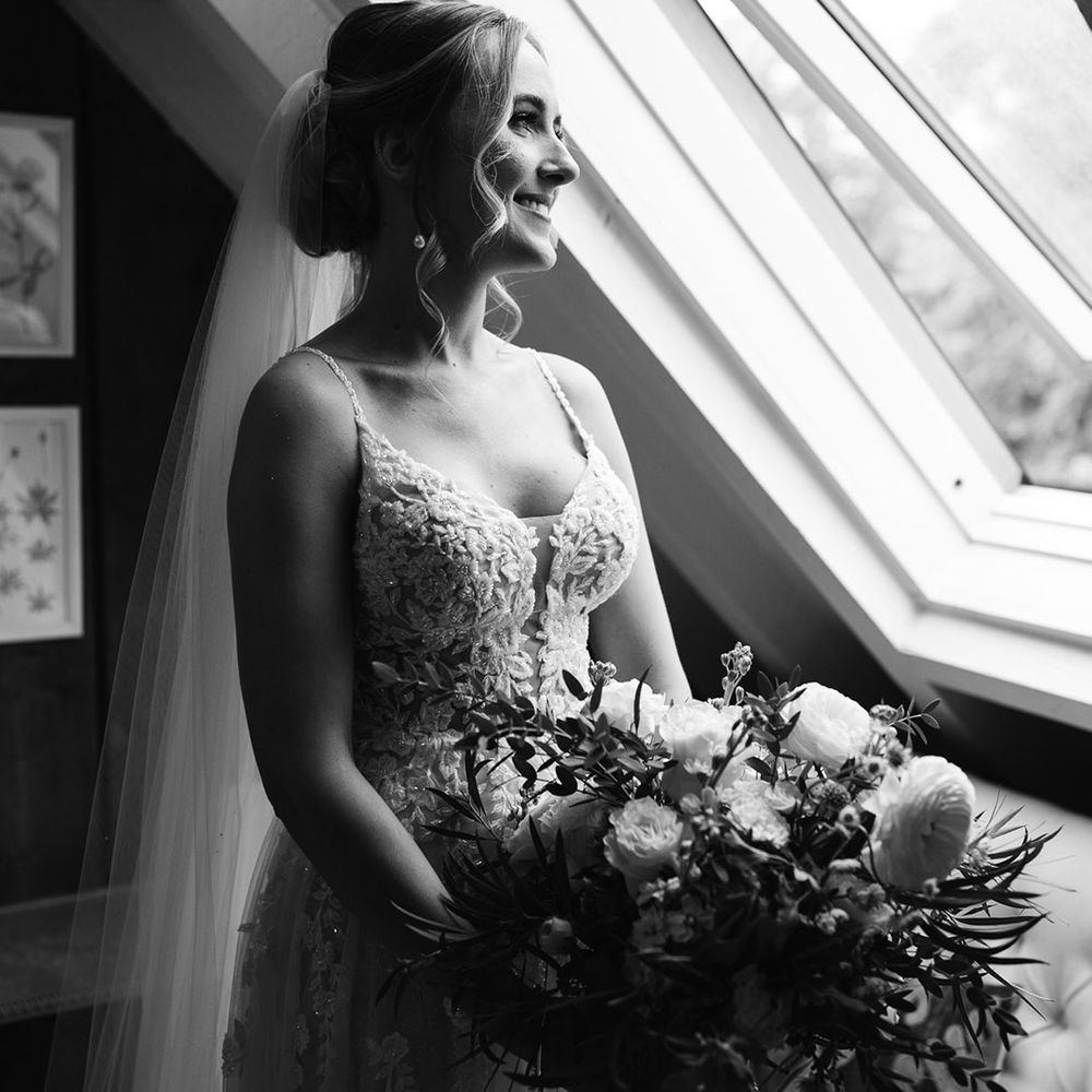 Bride in floral lace wedding dress looking out the window holding a white flower bouquet 