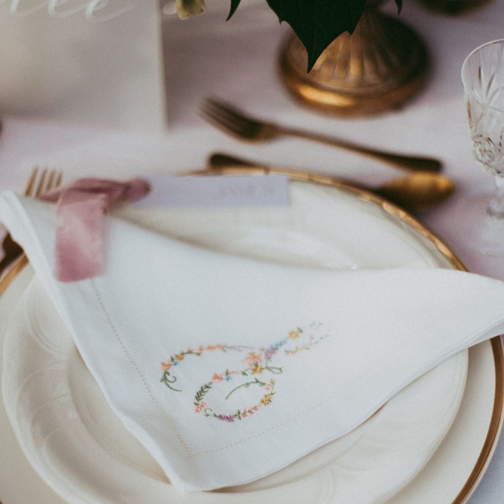 White delicate embroidered napkins with small colourful flowers and gold cutlery on neutral wedding tablescape