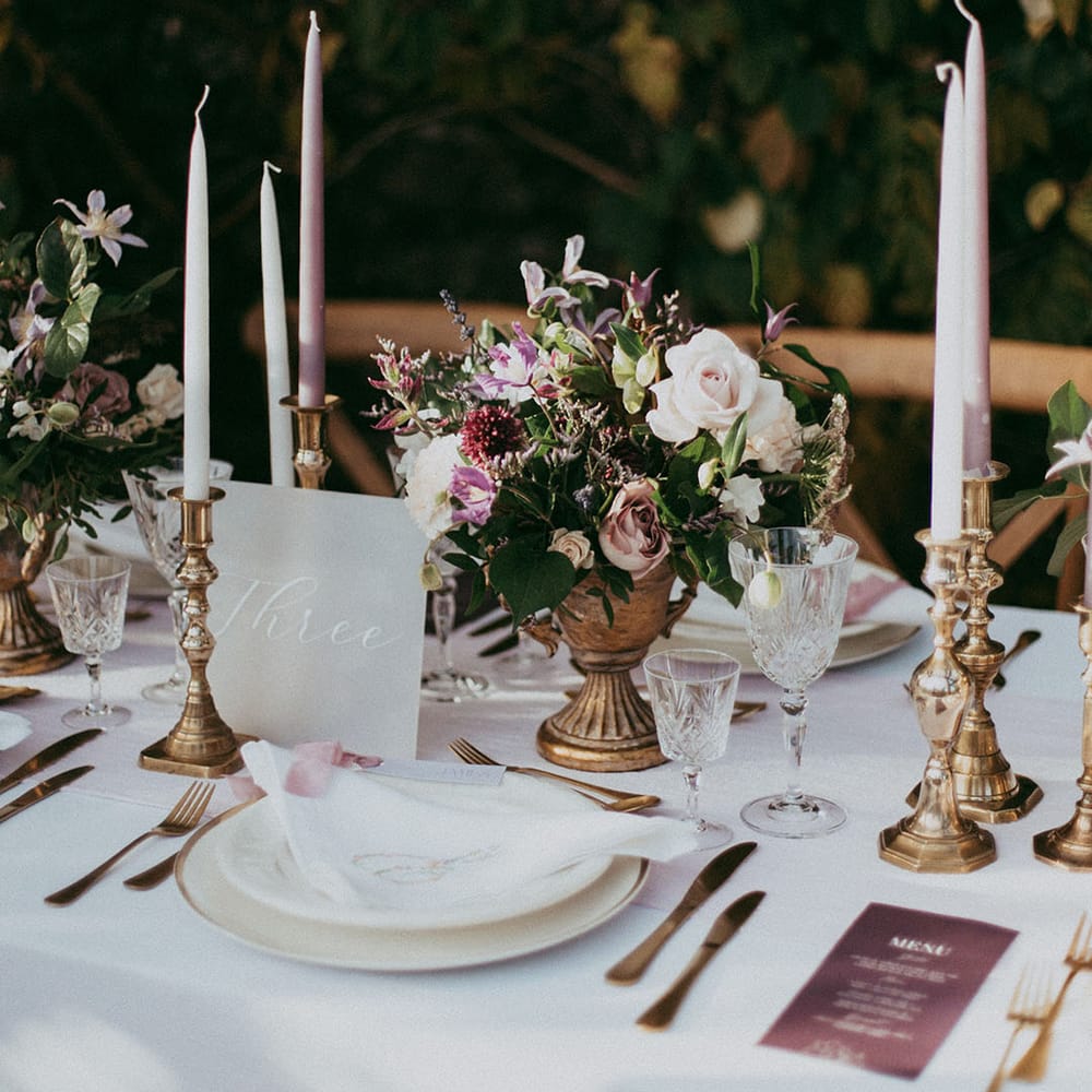Neutral, minimalistic wedding tablescape with white tapered candles, gold candle holders, mixed floral arrangements with white carnations, purple tulips, roses and foliage, and lavender wedding menus