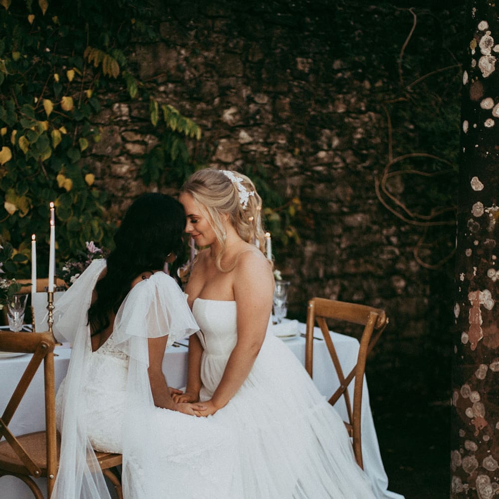 Bride in open back detail wedding dress with puff lace sleeves and bride in layered tulle sleeveless wedding dress with v neck detail holding hands at wedding reception