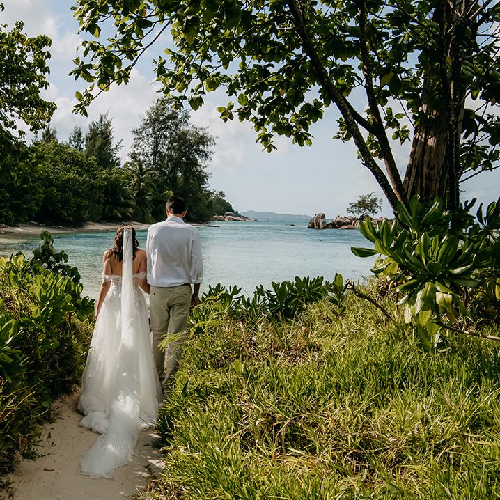 Bride in off the shoulder lace wedding dress with sweetheart neckline and lace floral details and cathedral length veil and groom in crisp white shirt, light coloured chinos, tan leather belt and white trainers walking through foliage on the beach