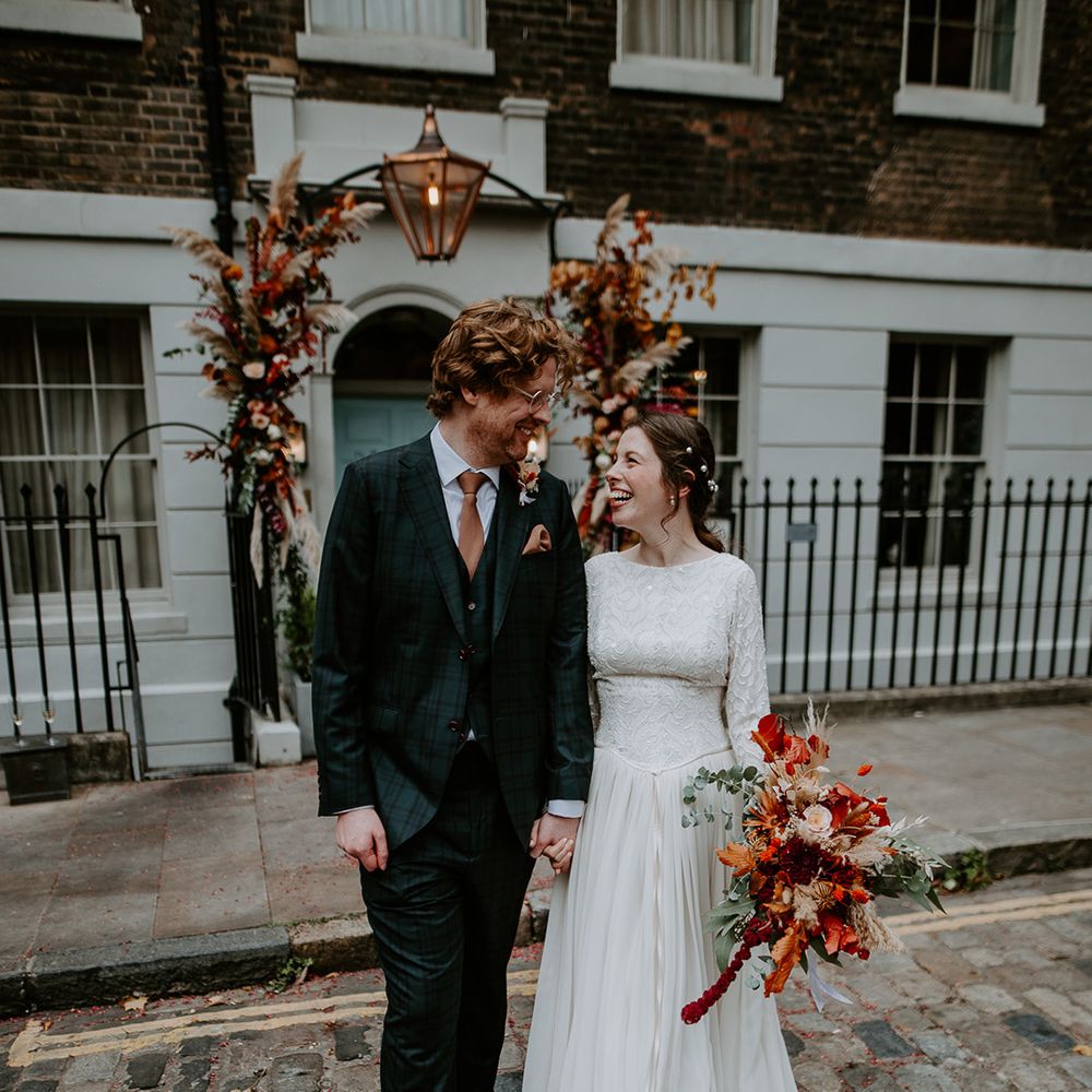 Groom in dark suit with orange tie, orange pocket square and autumnal boutonniere holding hands with bride in long sleeve lace vintage wedding dress and closed toe satin heels holding large autumnal bouquet with orange, black, green and yellow roses, peonies, foliage and pampas grass