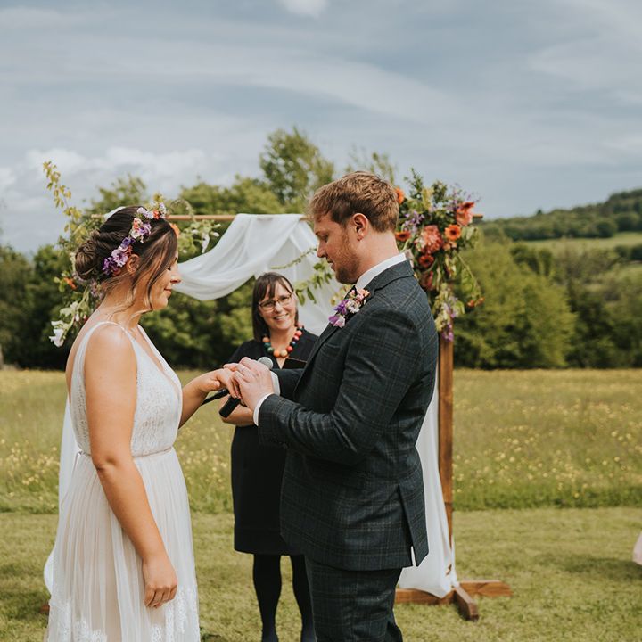 Groom places ring on his brides finger outdoors in front of wooden arch complete with white drapes and floral decor 