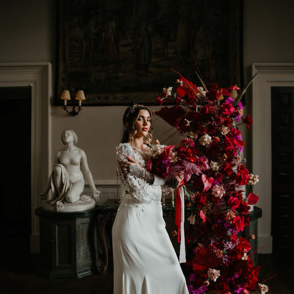 Bride standing by large standing floral decorations with hot pink, bright pink and viva magenta dahlias, orchids, roses, amaryllis, anthuriums and dried flowers wearing two piece bridal separates with puff sleeve sheer crop top with 3d appliqué flowers and full bodied silk skirt