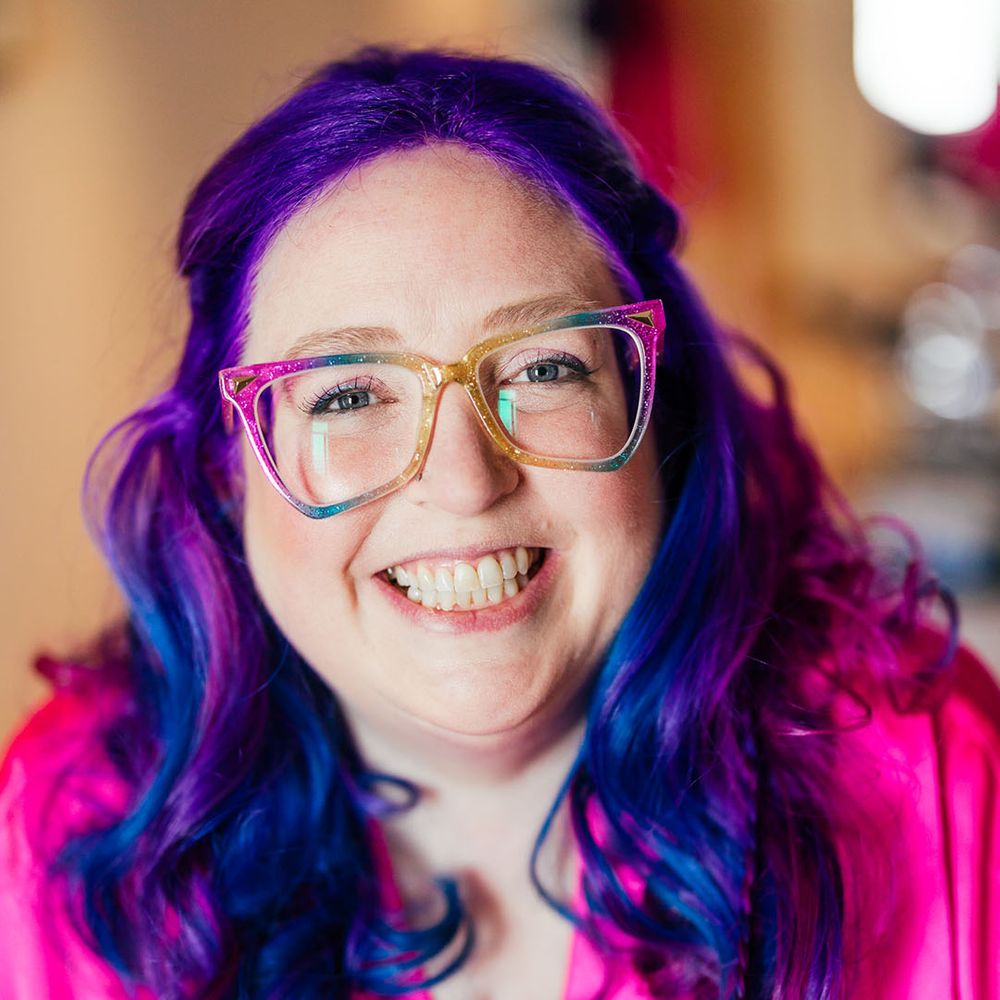 Bride with vibrant purple hair and purple cat eye glasses wearing bright pink getting ready robe before theme park wedding 