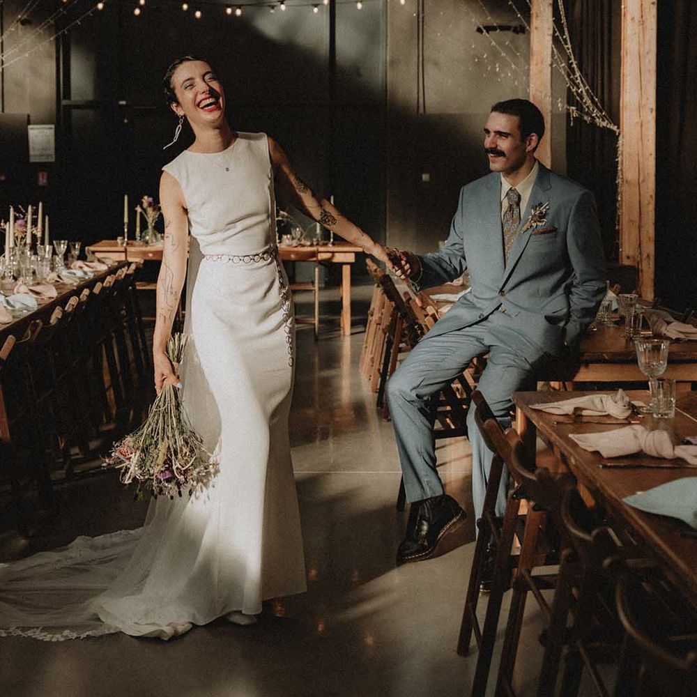 Lisa in pre loved fitted wedding dress laughs beside Gabriel in Gucci suit as festoon lighting hangs from above 