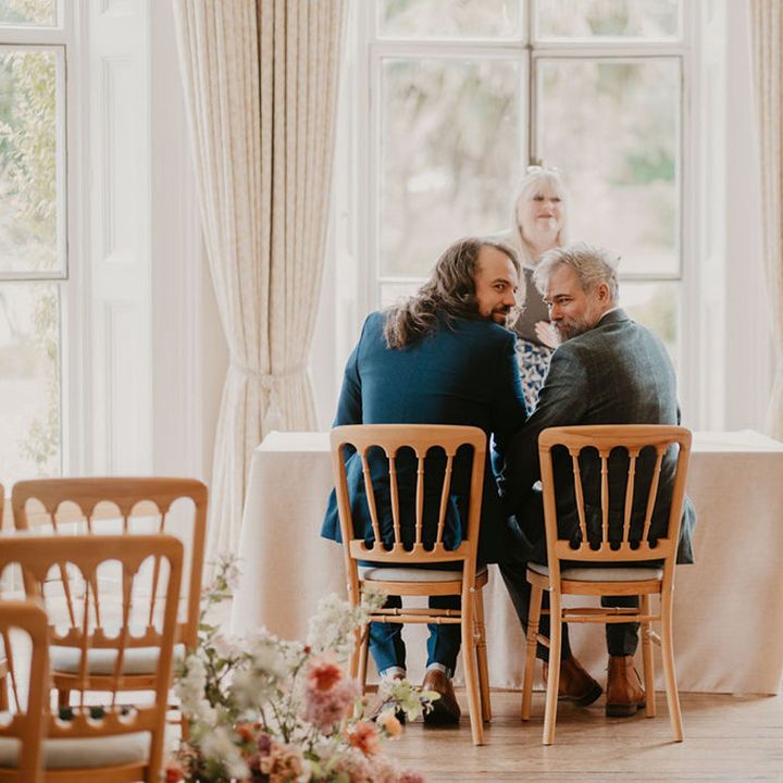 Grooms sit at table in front of window during wedding ceremony at the Cambridge Cottage 