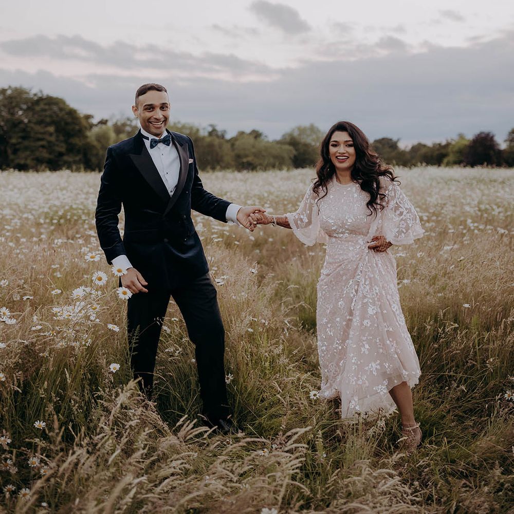 Bride in embellished long sleeve sheer dress stands beside groom in black velvet tuxedo standing in the grounds of Braxted Park wedding venues Essex