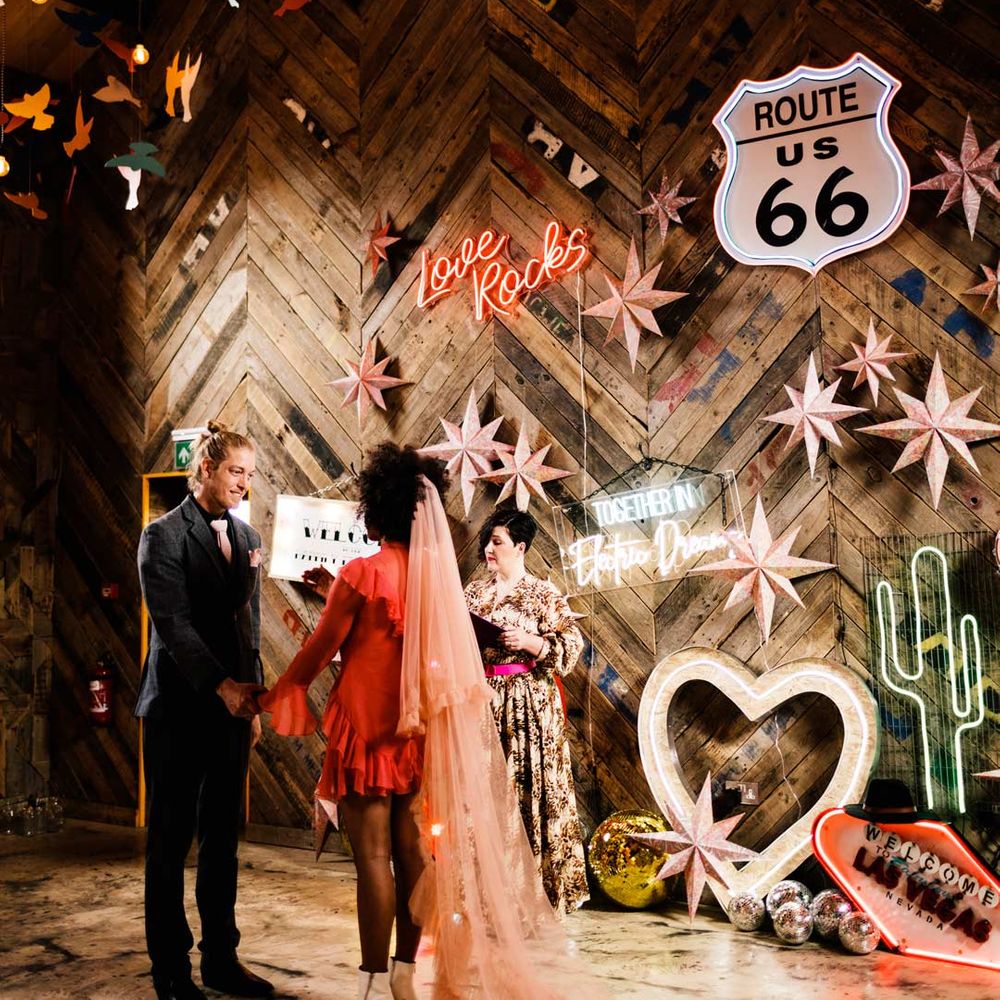 Bride in long sleeve short red wedding dress and pink two tier veil holding hands with groom in dark suit with an abundance of neon wedding signs 