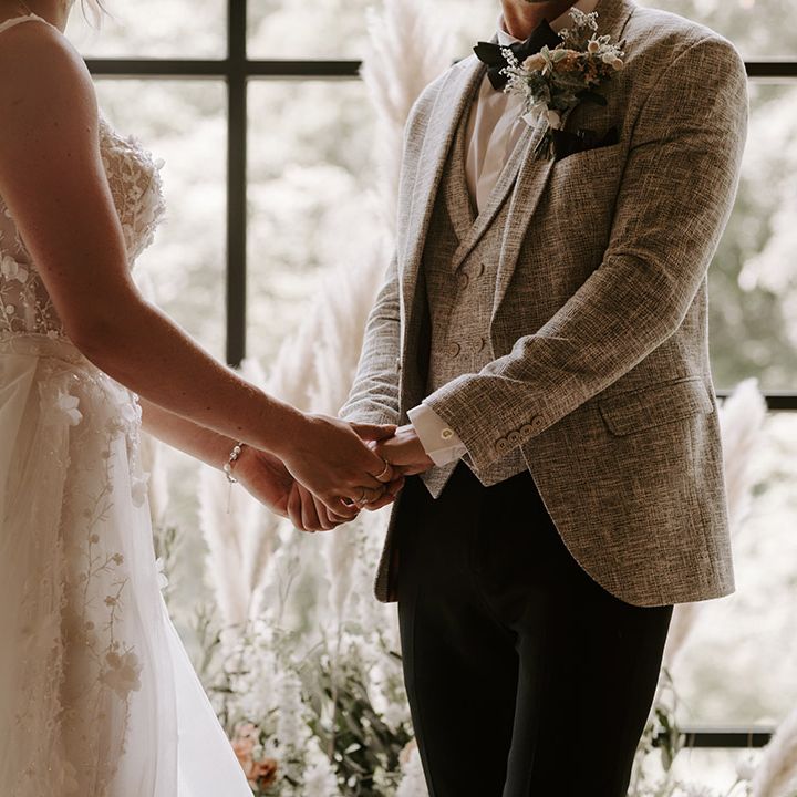 The bride in a floral applique Eleganza Sposa wedding dress and pearl veil holding hands with the groom in light grey suit at the ceremony 