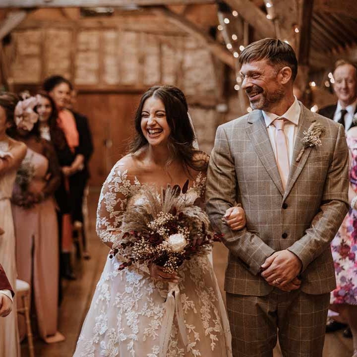 Bride in maroon bridal suit with grey waistcoat, dusky pink tie and autumnal dried flower boutonniere smiling at bride in long sleeve lace illusion sleeve wedding dress with overlay holding large autumnal boho dried flower bouquet 