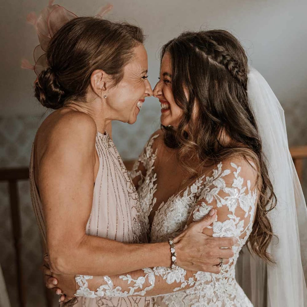 Bride in long sleeve lace illusion sleeve wedding dress with overlay hugging mother of the bride in dusky pink sleeveless dress and dusky pink fascinator 