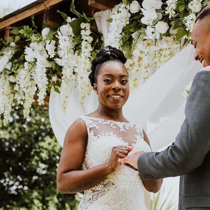 Bride in lace and mesh boatneck wedding dress with silver bridal hair accessories putting wedding ring on groom in charcoal grey grooms suit, light grey patterned waistcoat, light grey patterned tie and large white rose and foliage boutonniere 