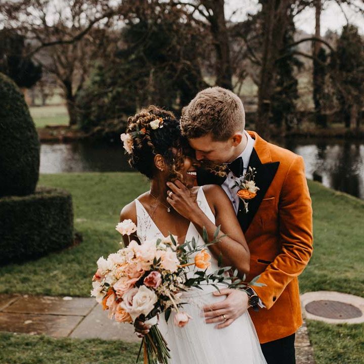 Groom in crushed burnt orange velvet grooms blazer, black bowtie and black suit trousers embracing bride in v neck sleeveless wedding dress with 3D floral applique train holding blush and orange garden roses, poppies and carnations, dried flowers and eucalyptus wedding bouquet at Brinsop Court wedding venue