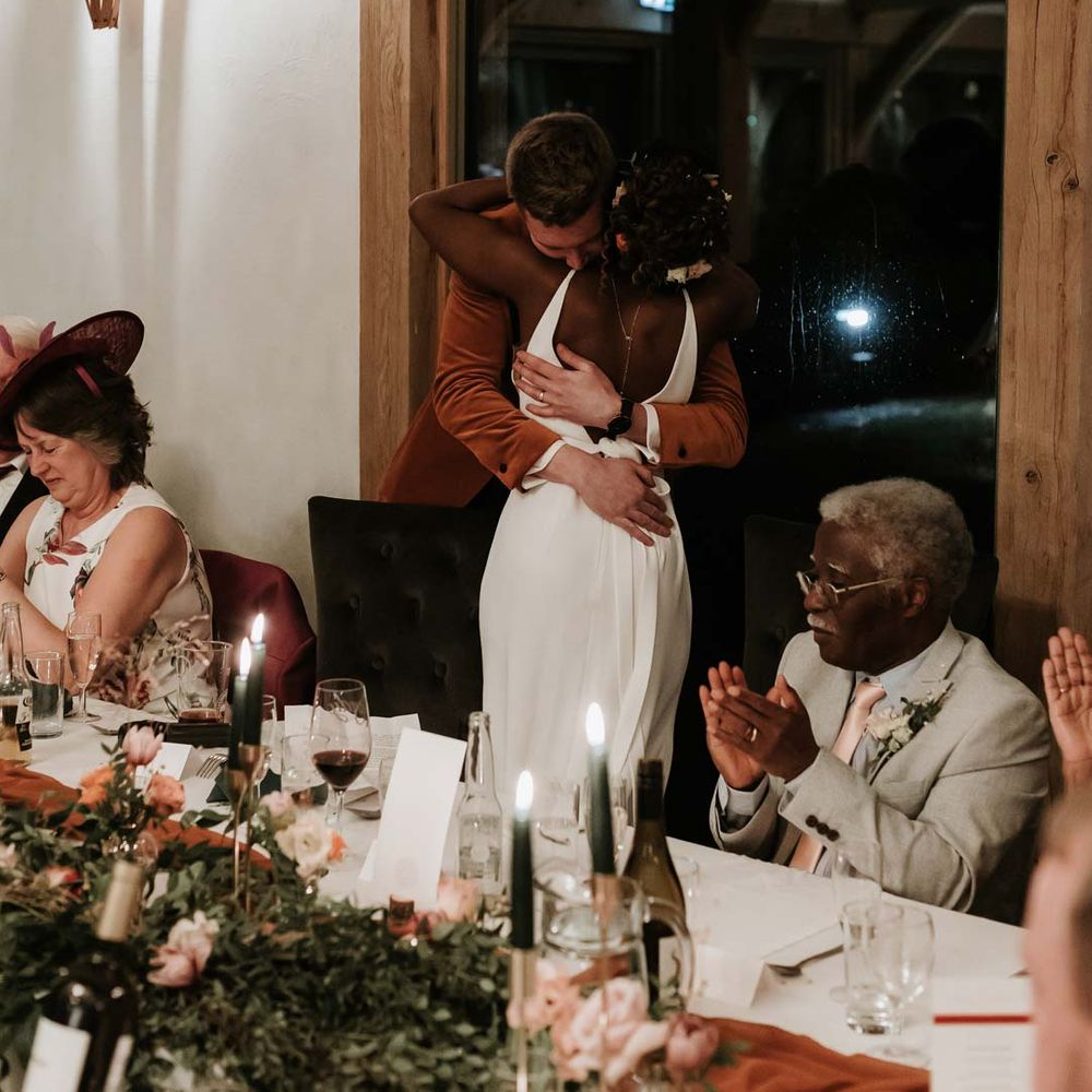 Bride in open back wedding dress with silver body chain hugging groom during wedding speeches