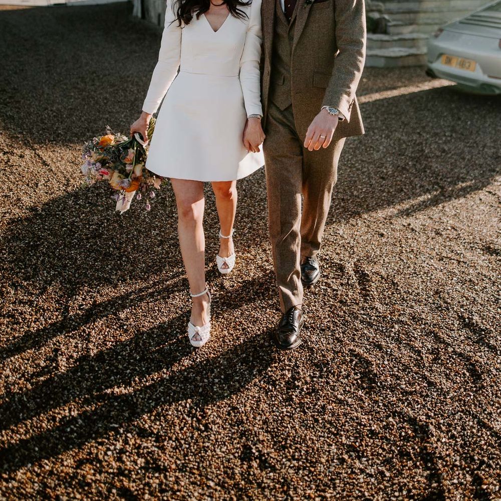 Groom in three piece chocolate brown grooms suit walking with bride in long sleeve short wedding dress, pearl bridal headband and platform pearl wedding heels at Willow Marsh Farm Loughborough 