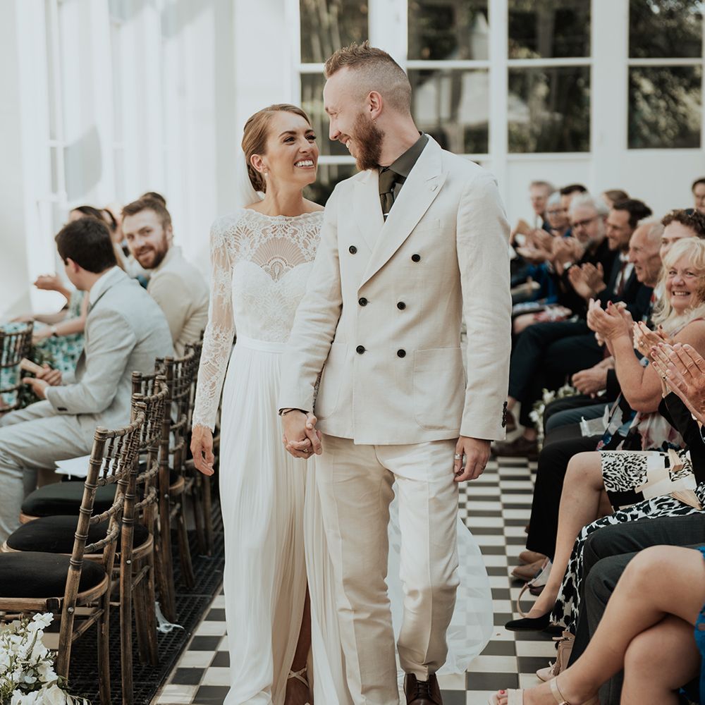 groom in a beige double breasted suit holding hands with his bride in a long sleeve wedding dress at Gunnersbury Park orangery wedding 