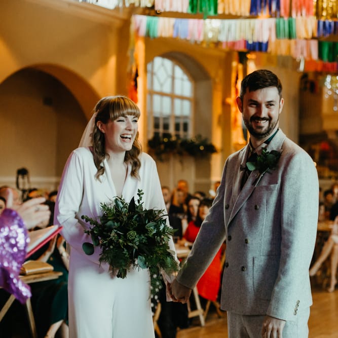 BRide in a jumpsuit holding a green foliage wedding bouquet smiling at the altar with her groom in a grey double breasted suit 