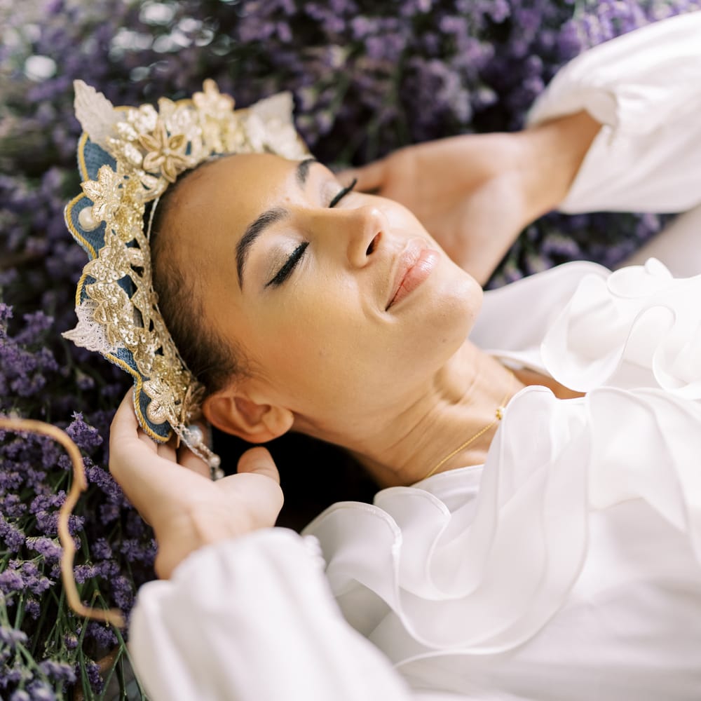 Bride lays back into lavender with eyes closed and holds golden embroidered crown