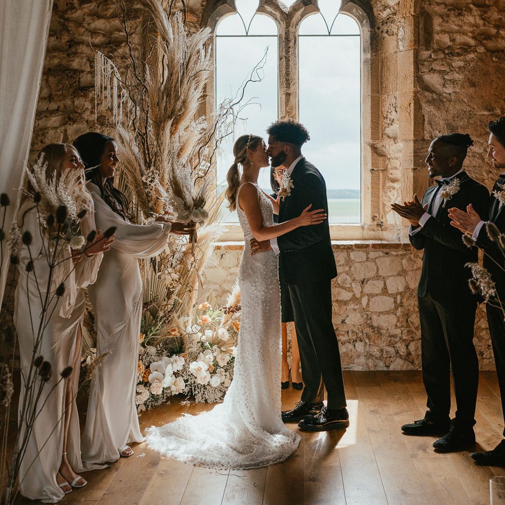 Groom in a tuxedo kissing his bride at the stone altar with window backdrop and dried and fresh flowers in neutral tones