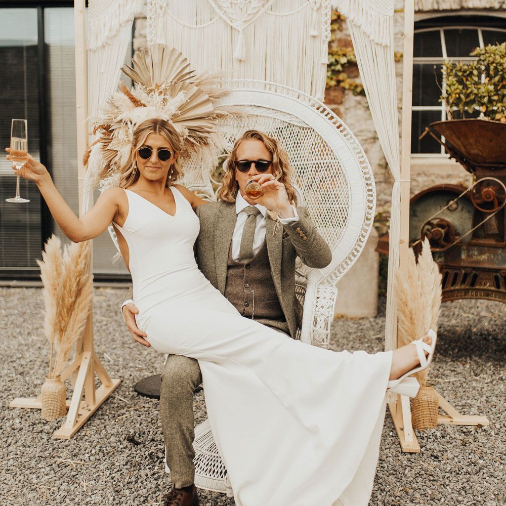 Bride in white Made With Love wedding dress and sunglasses holds up glass of sparkling wine as she sits on lap of groom in sage suit and sunglasses as he drinks beer whilst sat on white rattan throne in the courtyard of Anran Devon