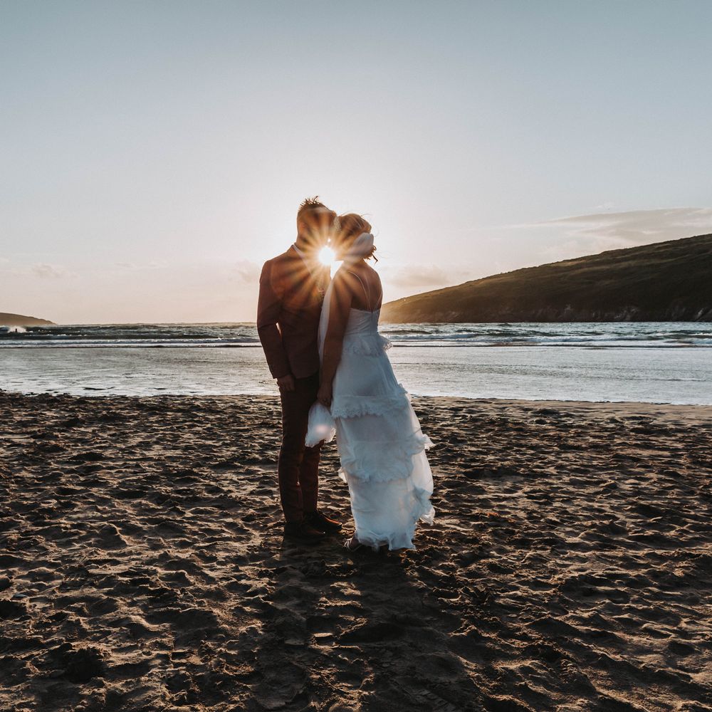 Bride & groom kiss as the sun sets behind them on the beach