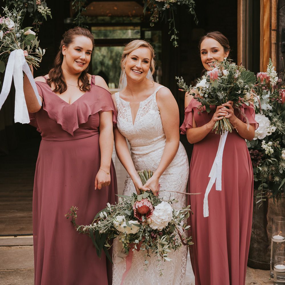 Bride in lace Justin Alexander wedding dress stands in between two bridesmaids in dusky pink cut out shoulder bridesmaid dresses all holding pink, white and green wedding bouquets at Tythe Barn wedding with barn wedding flowers
