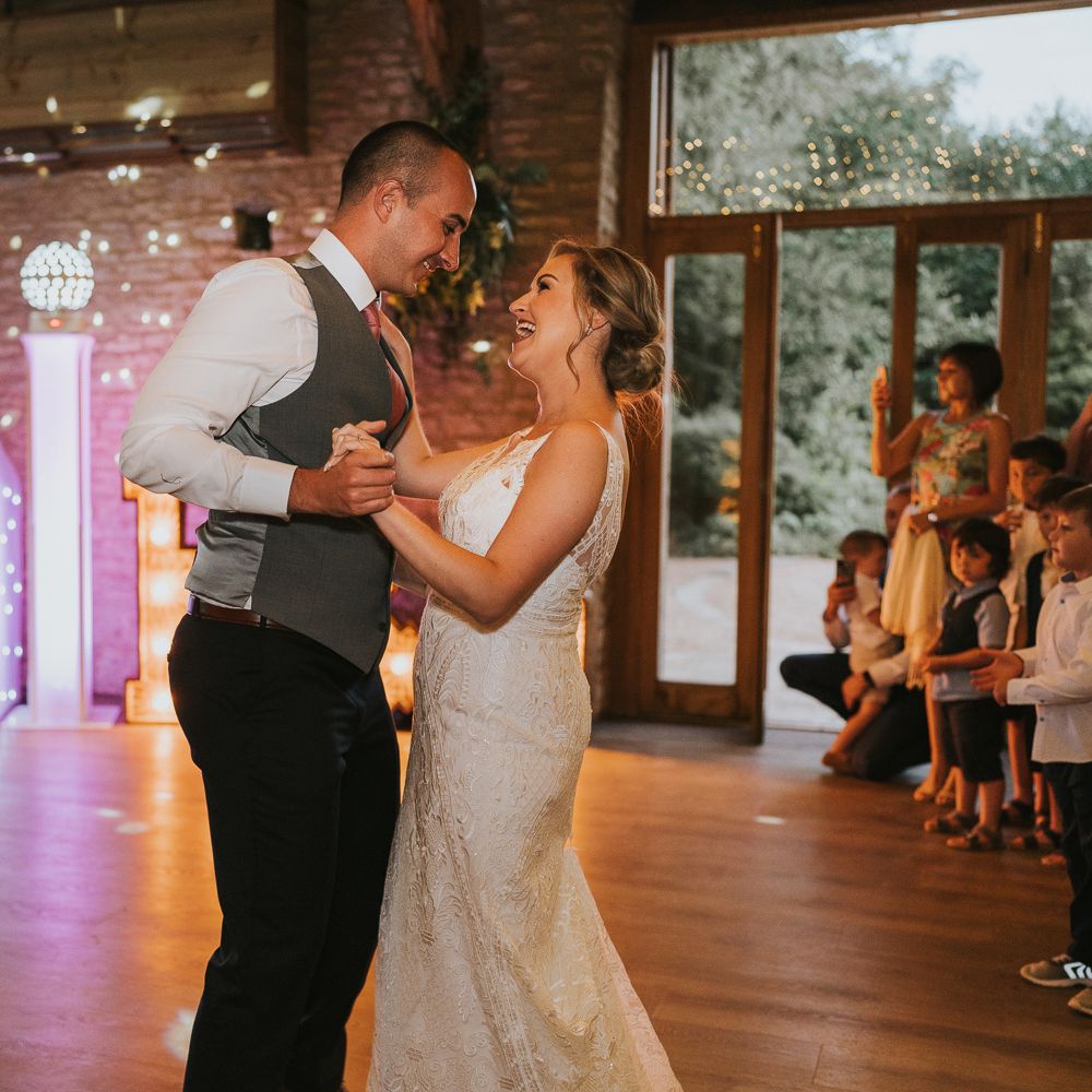 Bride in lace Justin Alexander Wedding dress and groom in white shirt, grey waistcoat and pink tie have their first dance at Tythe Barn wedding with barn wedding flowers