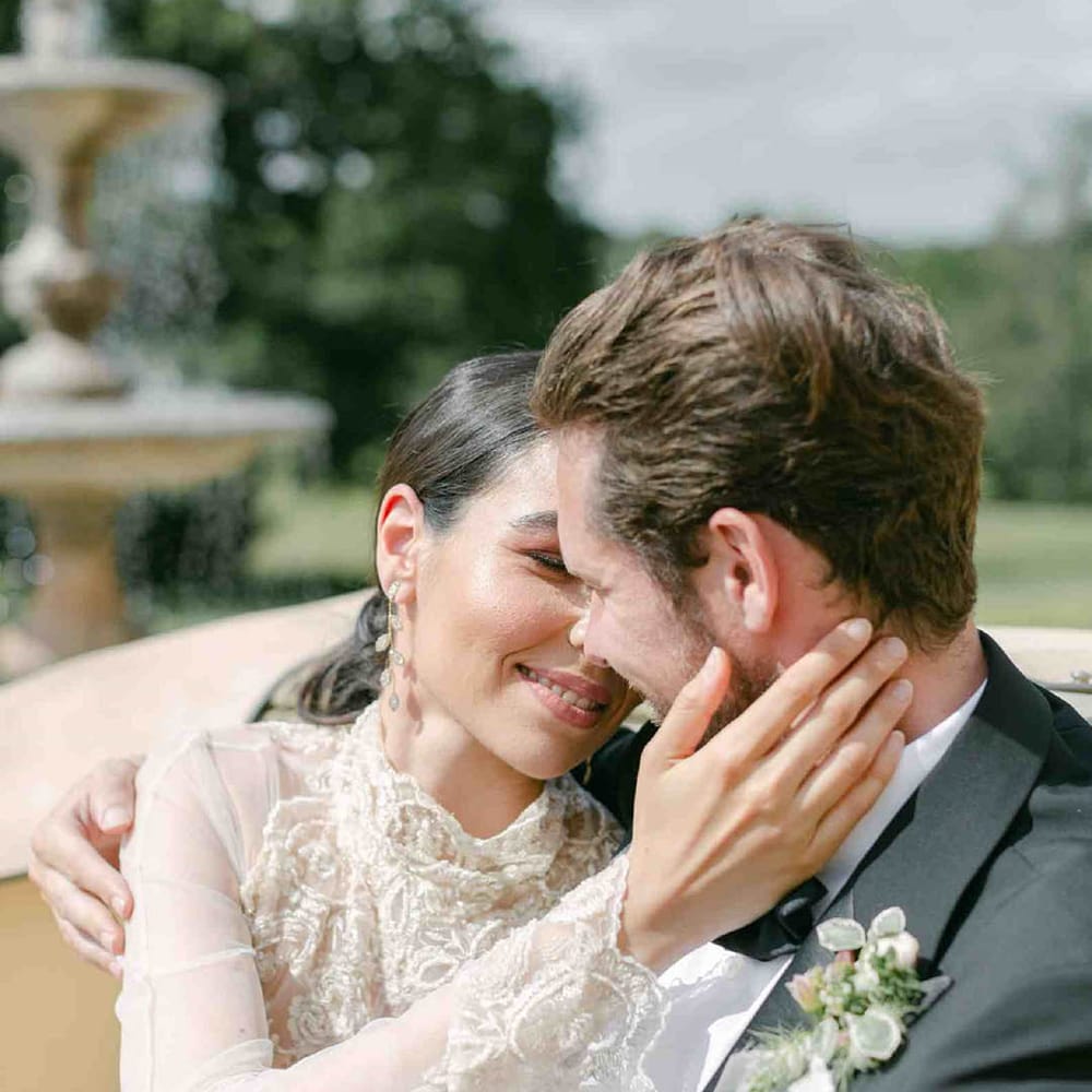 Bride in a sheer long sleeve wedding dress with high neck design and lace cuffs embracing her grooms face in the back of a vintage wedding car 