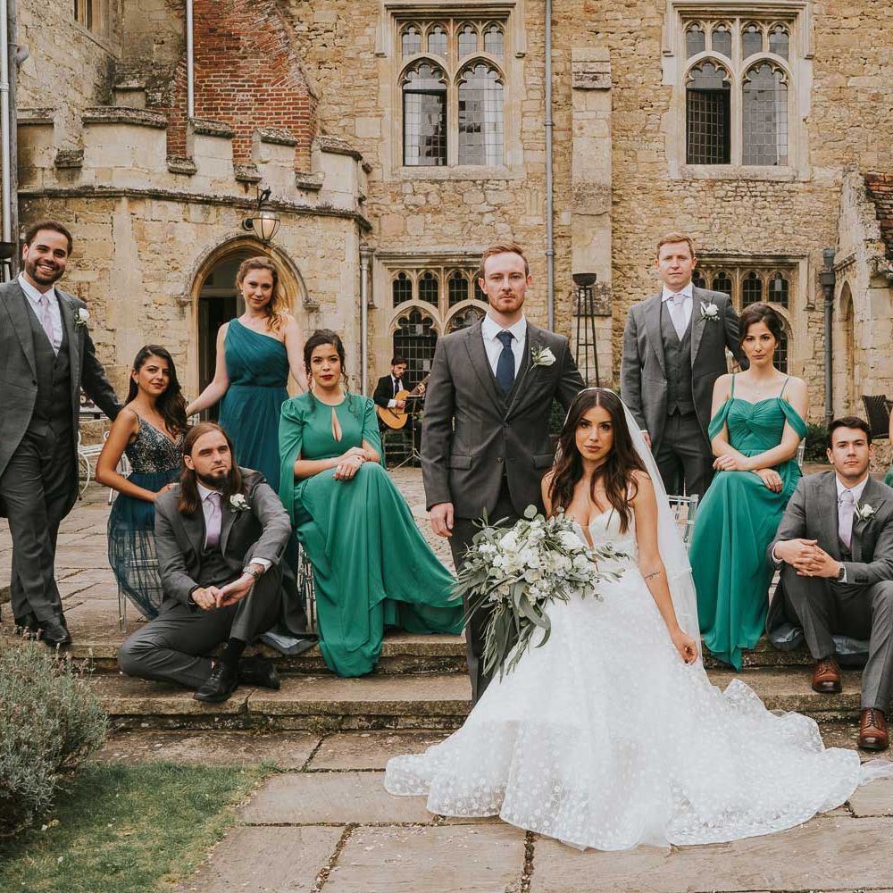 Bride in applique Pronovias wedding dress holding white and green bridal bouquet sits on step by standing groom in grey three piece suit surrounded by bridesmaids in assorted green bridesmaid dresses and groomsmen in grey morning suits in the grounds of Notley Abbey
