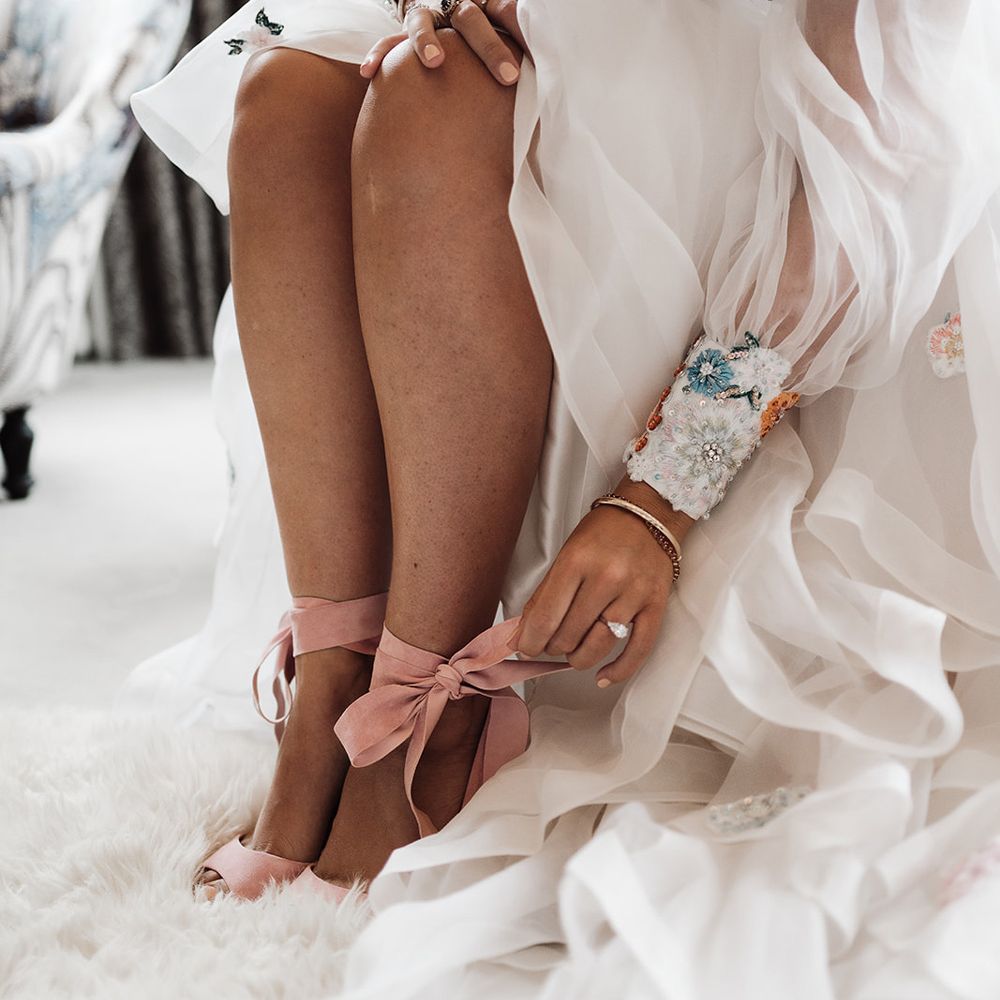 Bride in embroidered wedding dress sitting on stool ties bow on pink heels